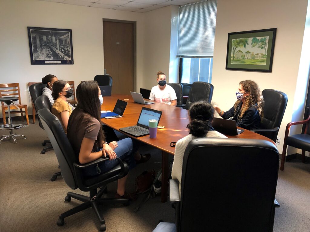 Group of Students in a room sitting around a large table having a discussion.